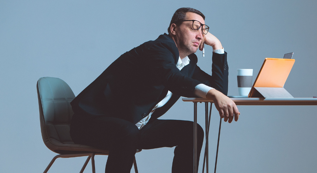 bored man in suit leaning on one hand, glasses askew, slouched at table in front of computer
