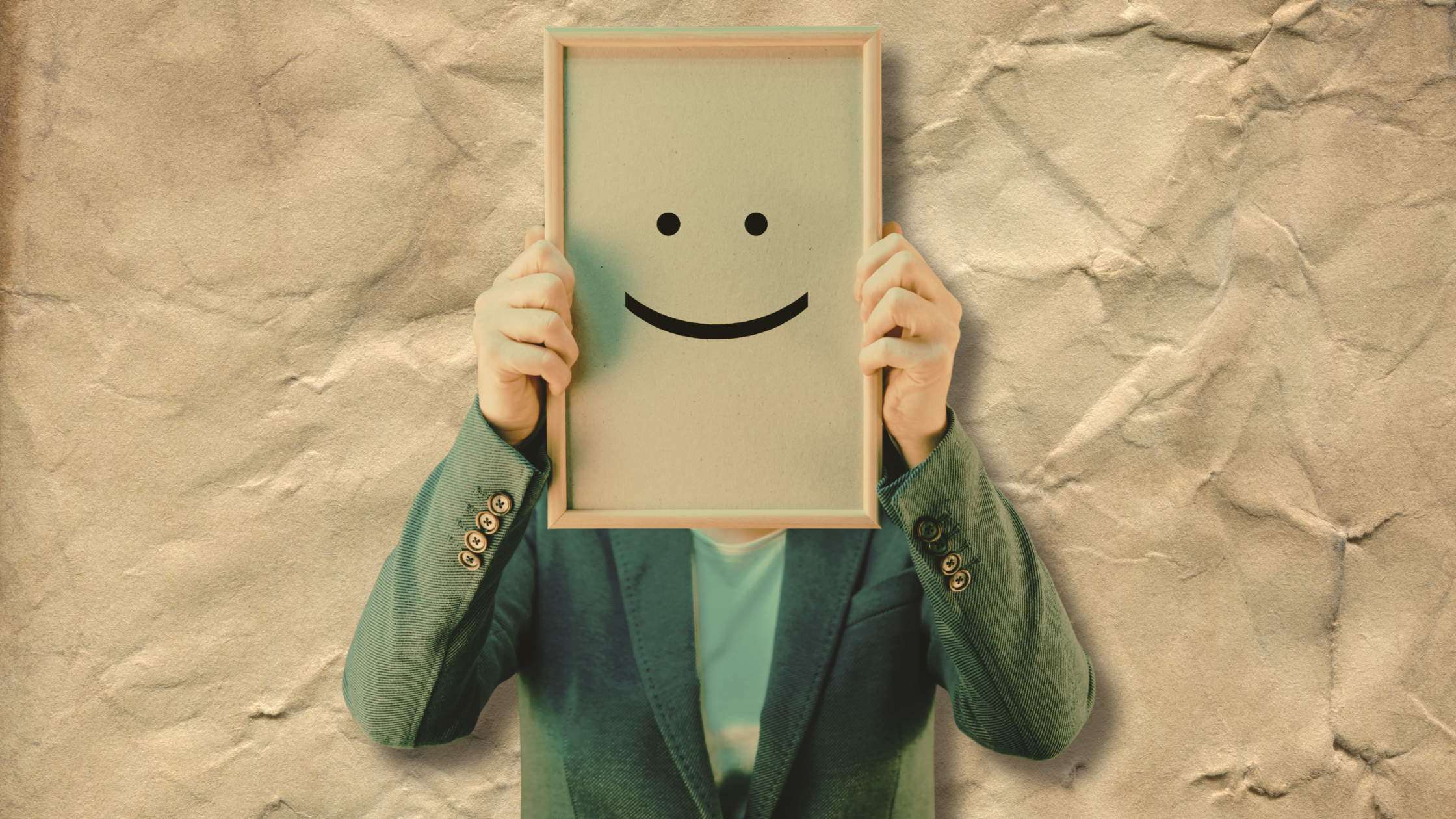 Man in suit jacket, holding up a board up with a smiley face in place of his own, on a background of crumpled paper